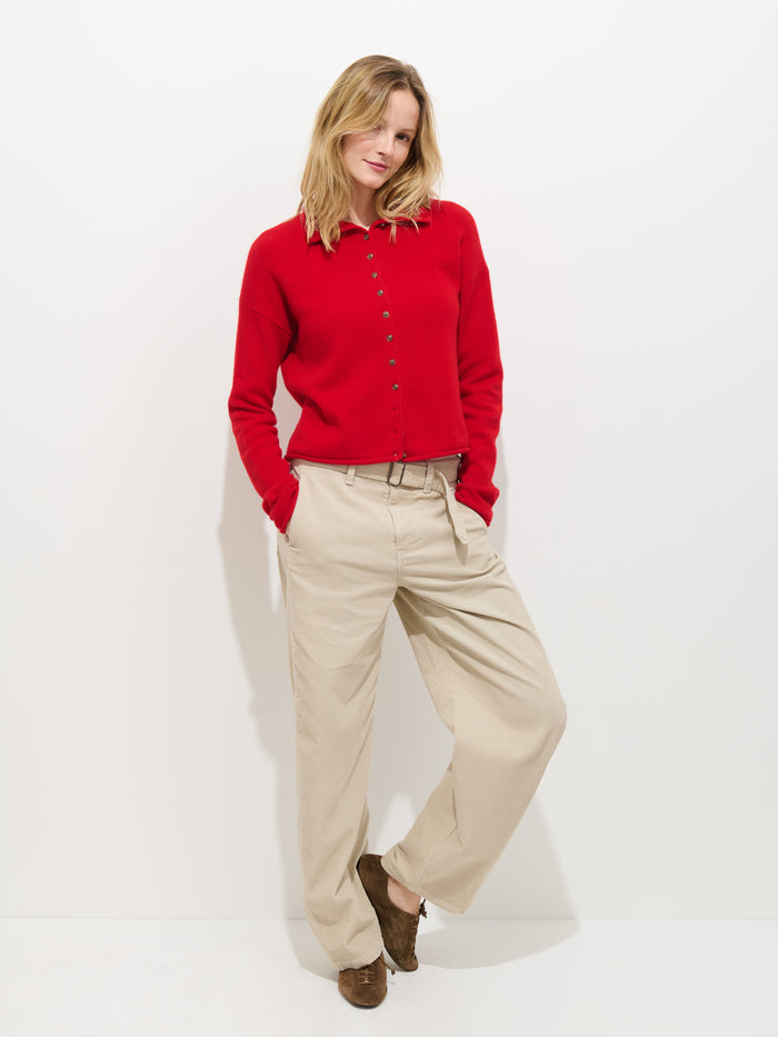 A person stands against a plain white background, wearing a red button-up sweater, the 9th Street Pant In Fine Wale Cord from Alex Mill in beige, and brown shoes, hands in pockets with a relaxed posture.