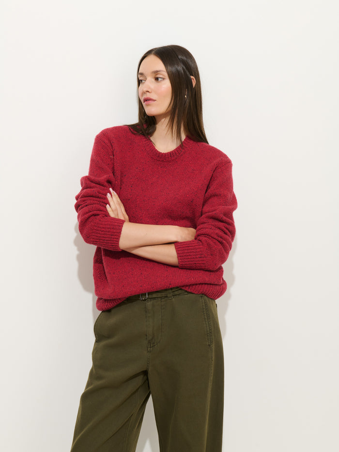A woman with straight dark hair stands against a white background, arms crossed and looking to the side, wearing olive green 9th Street Pant in pigment-dyed cotton and a loose red sweater.