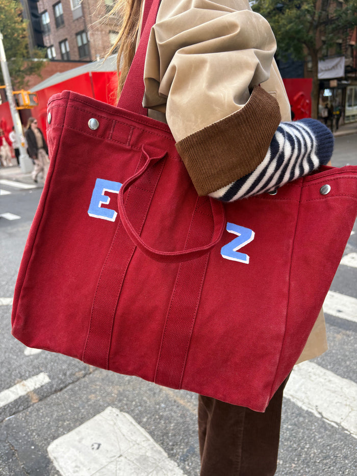 A person in a tan and brown coat with striped sleeves carries The Perfect Weekday Tote, a monogrammable cotton bag with blue and white letters E and Z, while crossing a city street.