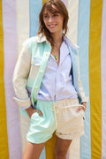A woman smiles in front of a pastel striped backdrop, wearing a light blue shirt, a mint and cream jacket, and matching Alex Mill x Quiet Town Saturday Shorts—small-batch canvas shorts made in Brooklyn—with her hands in her pockets.