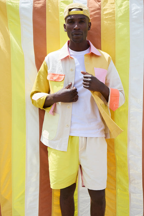 A man stands in front of striped yellow, orange, and white fabric, wearing a crisp white shirt, a yellow cap, and the Alex Mill x Quiet Town Saturday Shorts—crafted from patchwork shower curtain canvas as part of a small-batch collection made in Brooklyn.