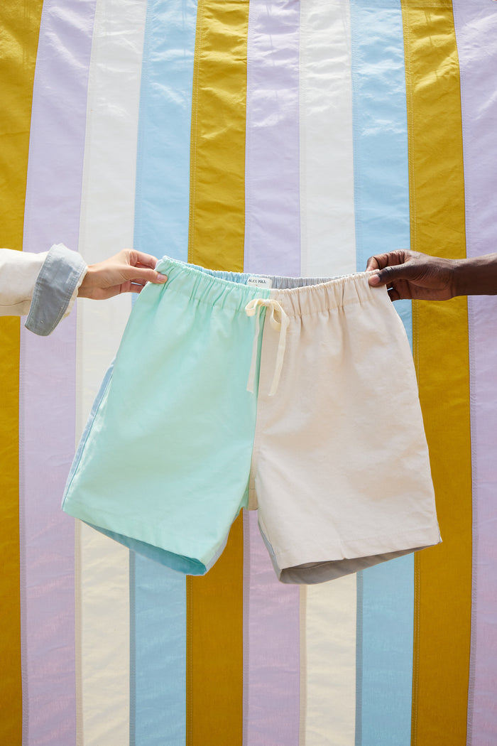 Two hands hold up the Alex Mill x Quiet Town Saturday Shorts—a small-batch, drawstring canvas style—framed by vertical yellow, blue, and white stripes. Crafted in Brooklyn, these pastel color-block shorts blend playful design with local artisanship.