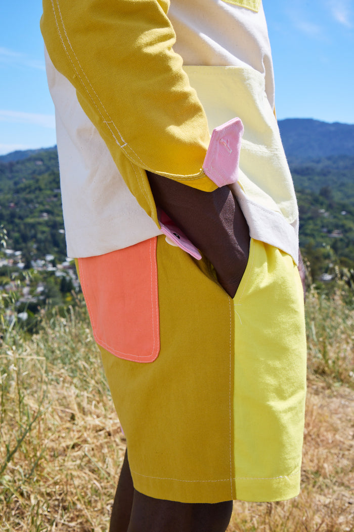 A person stands outdoors against a backdrop of hills and blue sky, wearing the Alex Mill x Quiet Town Saturday Shorts—crafted from color-blocked yellow, orange, white, and pink shower curtain canvas. The photo highlights only the lower torso and hand, showcasing this unique piece from a small-batch collection made in Brooklyn.