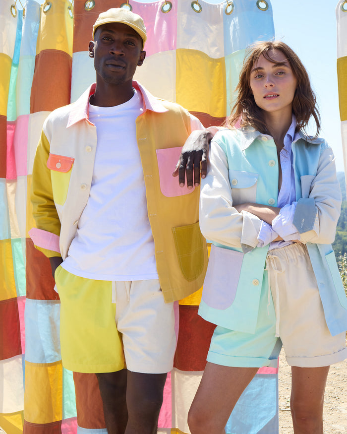 Two people stand in front of a colorful patchwork shower curtain canvas, wearing pastel and cream-colored casual outfits featuring the Alex Mill x Quiet Town Saturday Shorts with coordinating jackets. The person on the left wears a cap; both look confidently at the camera, perfectly embodying this small-batch collection made in Brooklyn.