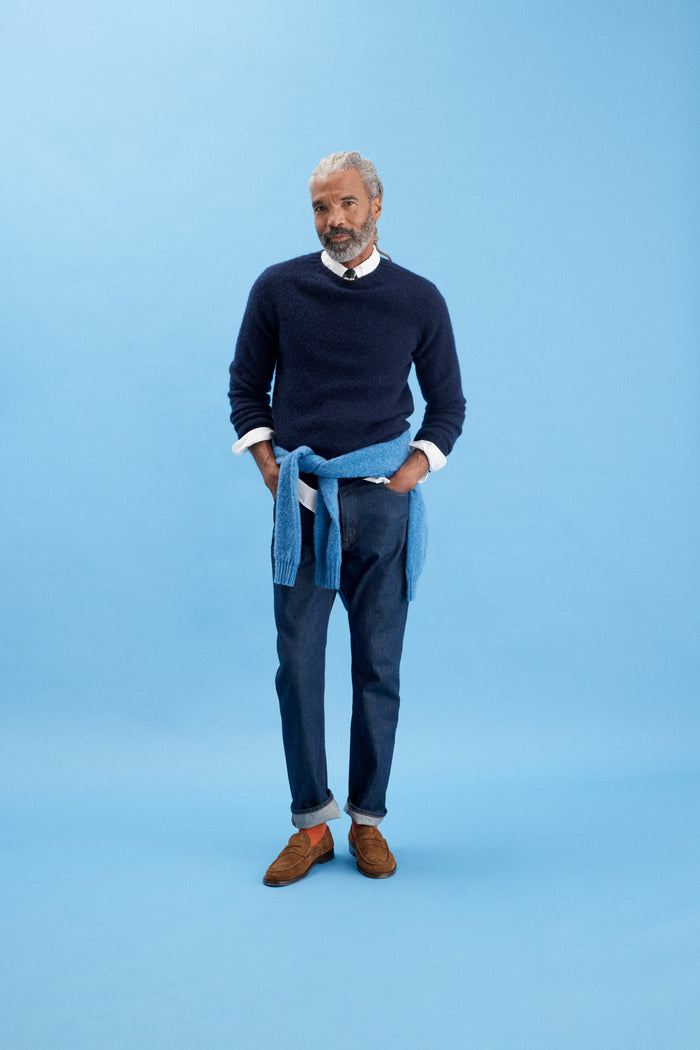 A man with gray hair stands against a blue background, wearing men’s sizing: a navy sweater over a white shirt, AM Original 5 Pocket Jean X J.Press, brown shoes, and a light blue sweater tied at his waist with hands in his pockets.