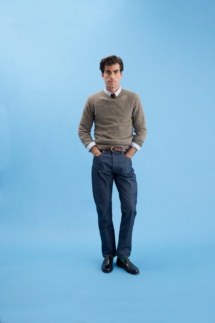 A man stands against a light blue background, wearing the AM Original 5 Pocket Jean X J.Press—100% cotton pants styled with a tan sweater, white shirt, and red tie. Hands in pockets, he looks straight ahead in this classic collaborative look.