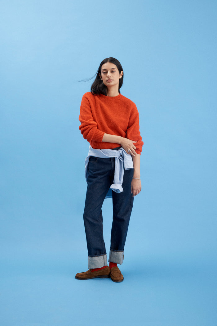 A woman with long dark hair and a relaxed expression stands against a blue background in a bright orange sweater, brown loafers, a light blue shirt tied at her waist, and AM Original 5 Pocket Jean X J.Press dark cuffed 100% cotton pants.