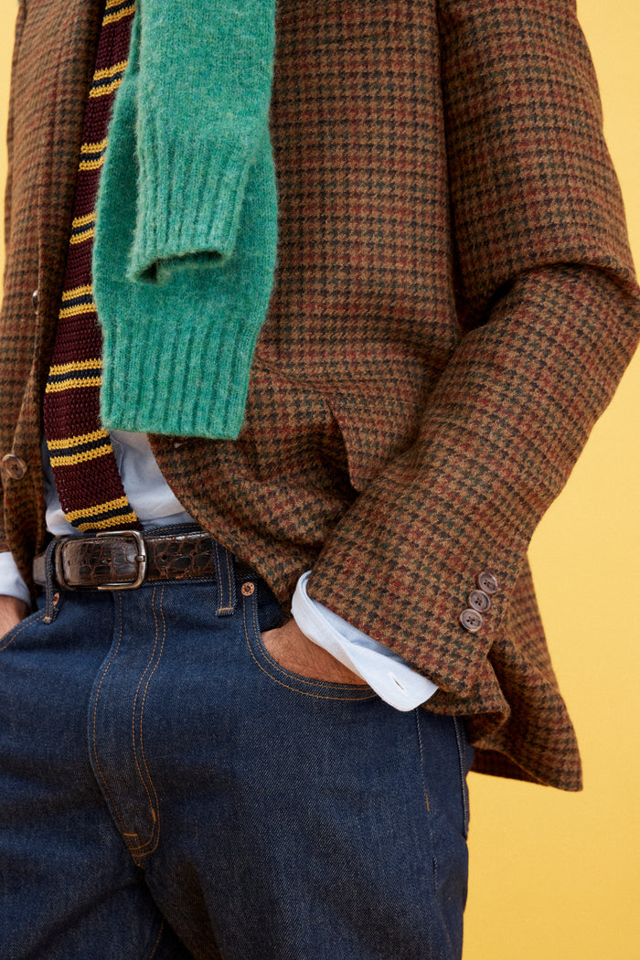 A person wears a brown plaid blazer, a striped tie, a white shirt, and an AM Original 5 Pocket Jean X J.Press in men's sizing, with a green sweater over their shoulders, standing against a yellow background with hands in jean pockets.