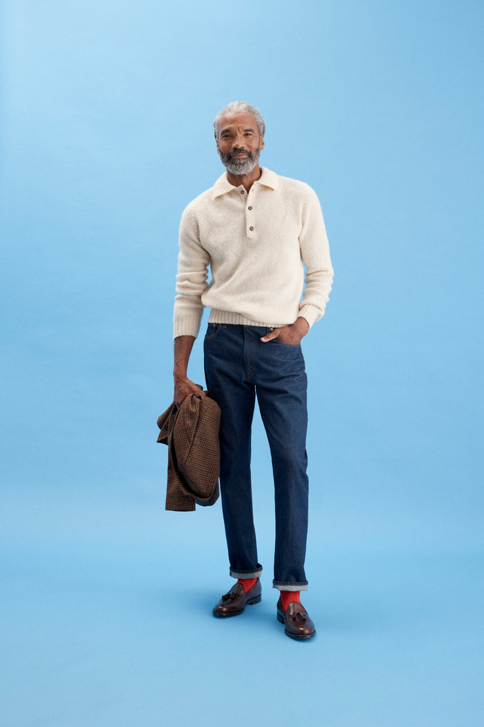 A man with gray hair and a beard stands against a light blue background. He wears a cream sweater, AM Original 5 Pocket Jean X J.Press cotton pants, brown loafers with red socks, and holds a brown jacket in his left hand.