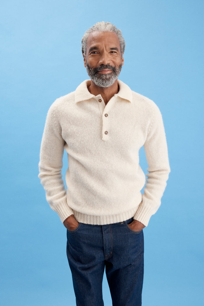 A man with gray hair and a beard stands against a light blue background, wearing a cream sweater with buttons and dark blue AM Original 5 Pocket Jean X J.Press in men's sizing. He has his hands in his pockets and is smiling slightly.