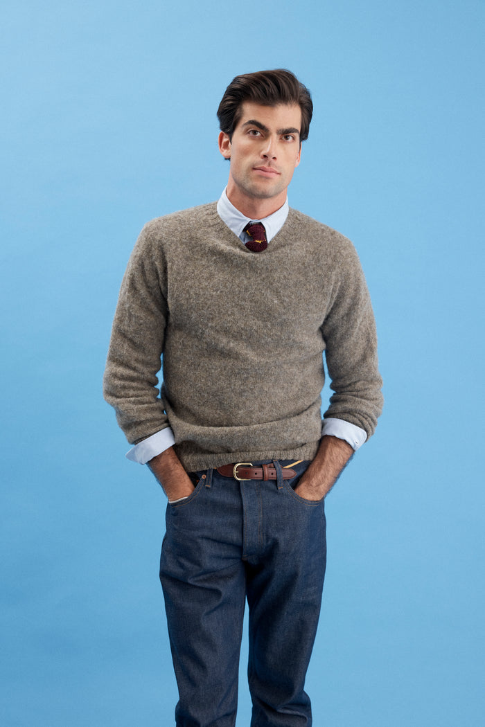 A man stands against a light blue background, wearing the AM Original 5 Pocket Jean X J.Press in blue 100% cotton, paired with a brown sweater layered over a collared shirt and tie. His hands are in his pockets as he looks at the camera neutrally.