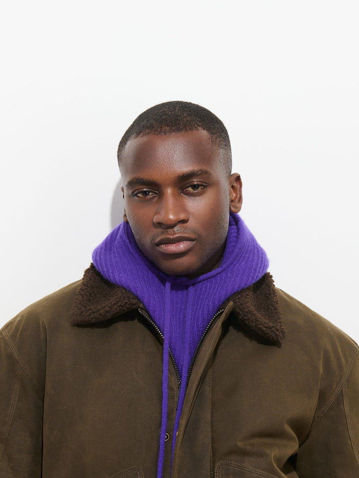 A man with short hair stands against a plain white background, wearing a brown jacket with a shearling collar and a bright purple scarf, effortlessly paired with the luxurious Balaclava in Cashmere.