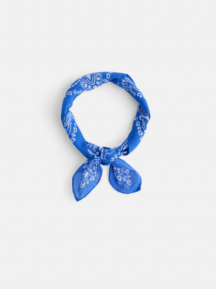 The Bandana in Cotton Voile, featuring a blue hue with white paisley patterns and crafted from soft cotton voile, is tied in a loose knot and displayed on a plain white background.