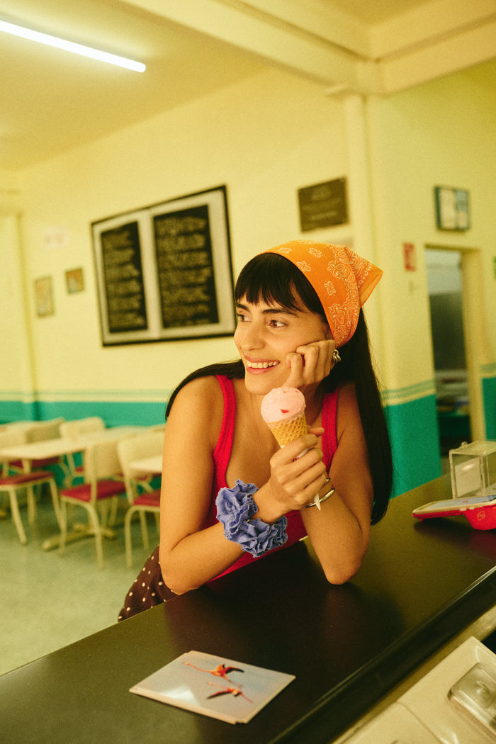A woman smiles while holding an ice cream cone, leaning on a counter in a retro-style café with green and cream walls, wearing the Bandana in Cotton Voile and a red tank top.
