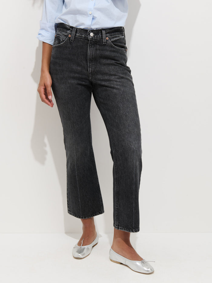 A person in Bessette Mid Rise Boot Cut Jeans, a light blue long-sleeve shirt with rolled sleeves, and silver ballet flats stands against a plain white background.