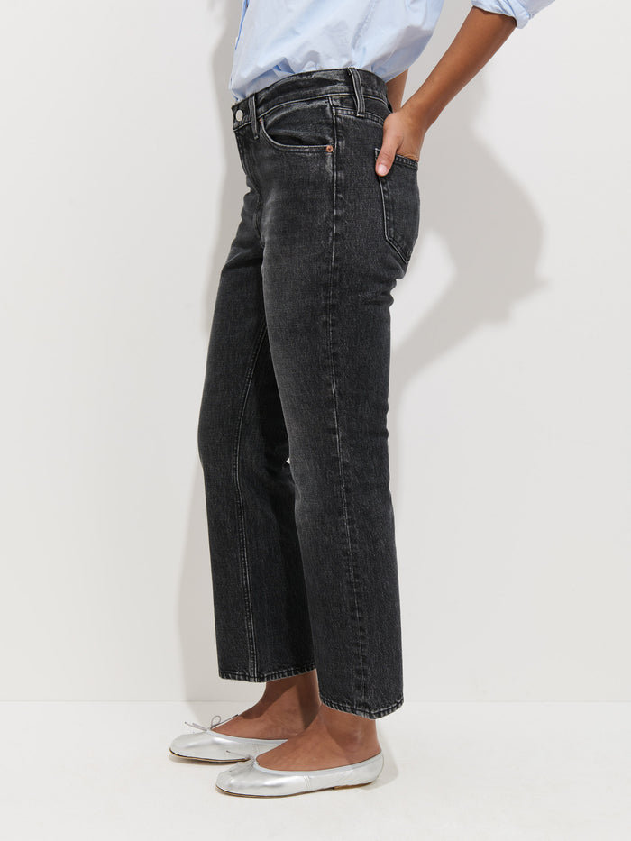 Only the lower half of a person is shown, standing against a white background, wearing Bessette Mid Rise Boot Cut Jeans in black with a light blue rolled-sleeve shirt and silver ballet flats.