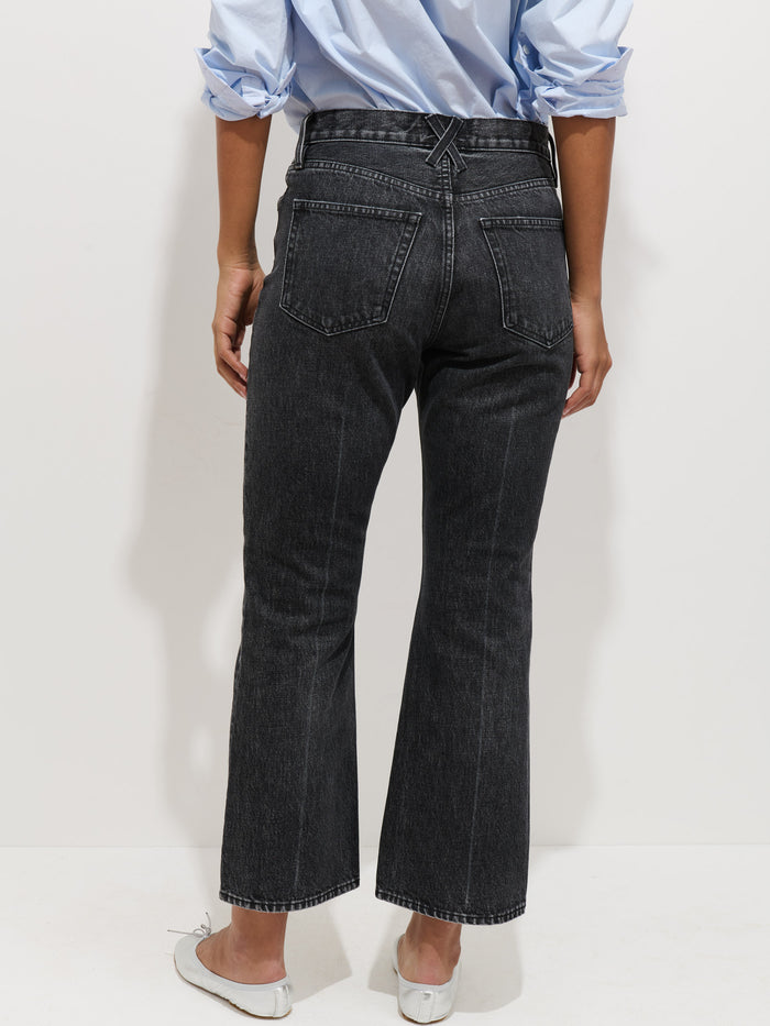 A person stands with their back to the camera, wearing a light blue shirt with rolled-up sleeves, Bessette Mid Rise Boot Cut Jeans in black 100% cotton, and white flats against a plain light background.