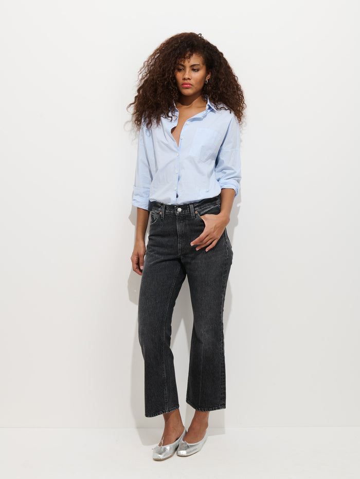 A woman with curly hair wears a light blue button-up shirt, silver shoes, and the Bessette Mid Rise Boot Cut Jean, standing against a plain white background.
