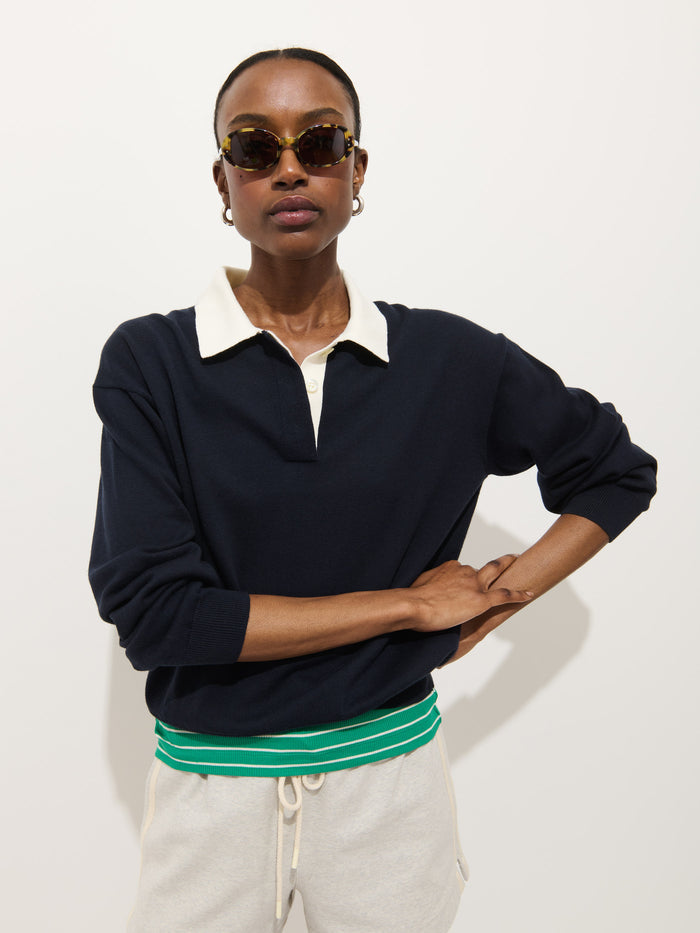 A woman wearing sunglasses and the Bond St Polo in Cotton Cashmere, navy with a white collar, paired with light gray sweatpants featuring a green and white striped waistband, stands against a plain white background.