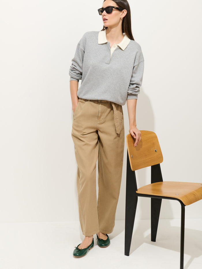 A woman wearing sunglasses and the Bond St Polo In Cotton Cashmere (a relaxed fit gray collared sweater), beige wide-leg pants, and green flats stands with one hand in her pocket, leaning on a wooden chair against a white background.