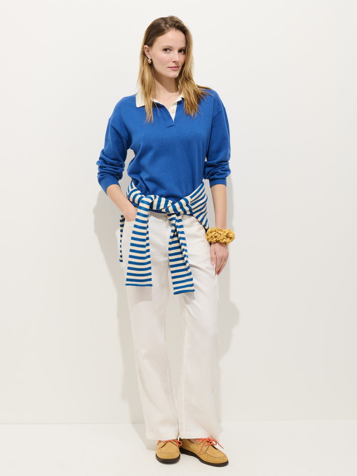 A woman stands against a white background wearing the Bond St Polo In Cotton Cashmere in rugby blue, paired with white pants, tan shoes, a blue-and-white striped sweater tied around her waist, and a scrunchie on her wrist.