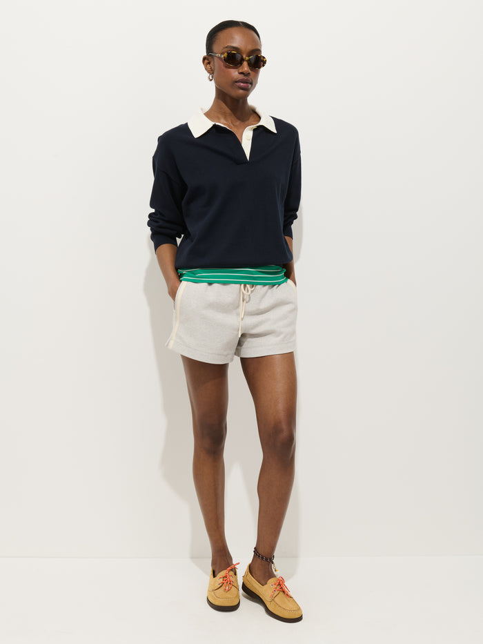 A person stands against a plain white background with hands in their pockets, wearing the Bond St Polo In Cotton Cashmere, layered over a green top with beige drawstring shorts, tan shoes, and sunglasses.
