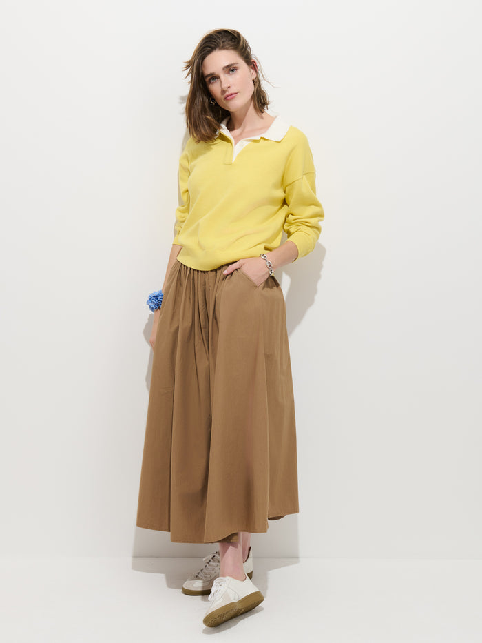 A person stands against a white wall wearing the Bond St Polo in Cotton Cashmere with a white collar, paired with a brown midi skirt, white sneakers, and a blue bracelet. Their hands are tucked into the skirt pockets.