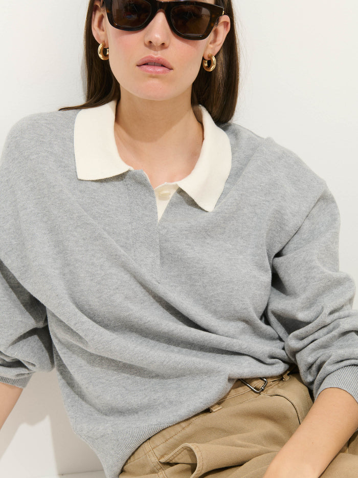 A woman wearing dark sunglasses, gold hoop earrings, and the Bond St Polo In Cotton Cashmere with a white collar and beige pants sits against a white background.