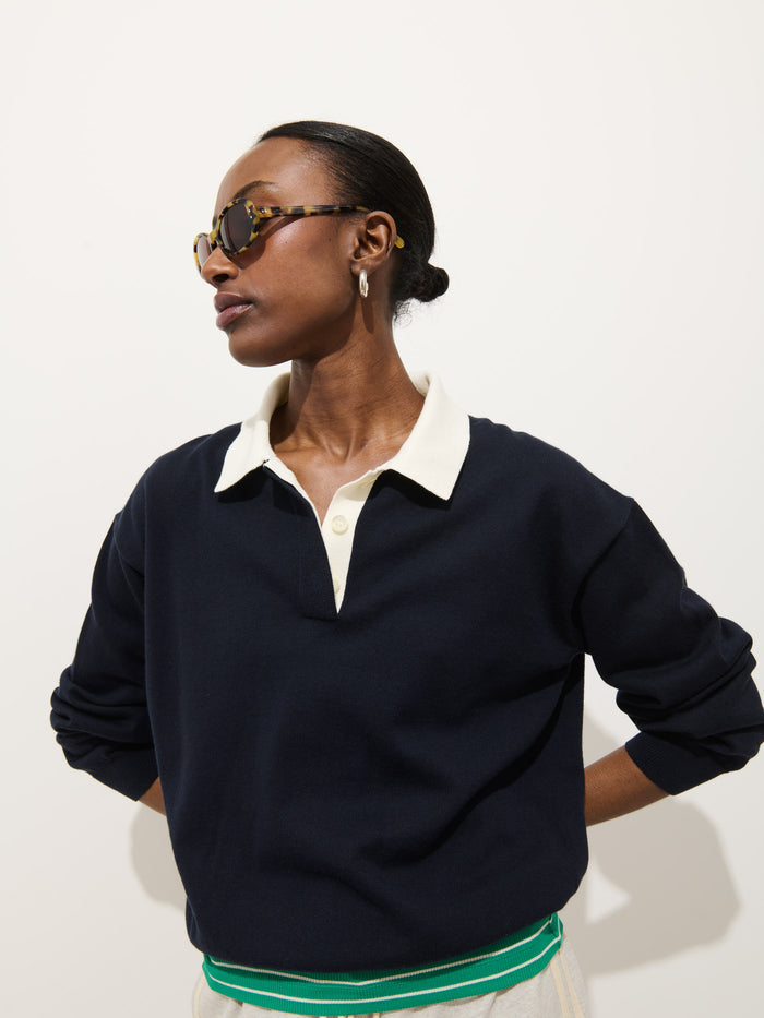 A person wearing sunglasses and the Bond St Polo In Cotton Cashmere, paired with cream pants with green and white stripes, stands against a plain white background with their hands behind their back.