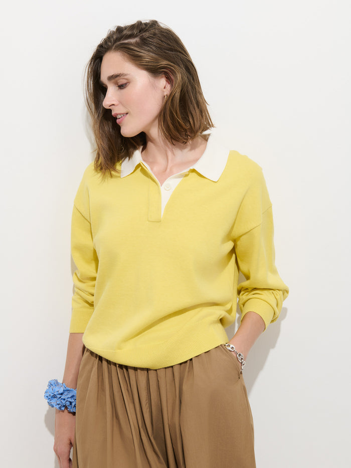 A woman with shoulder-length brown hair stands against a white background, looking to her left with one hand in her pocket and a blue scrunchie on her wrist. She wears the Bond St Polo In Cotton Cashmere—a relaxed fit, collared yellow sweater—paired with a tan skirt.