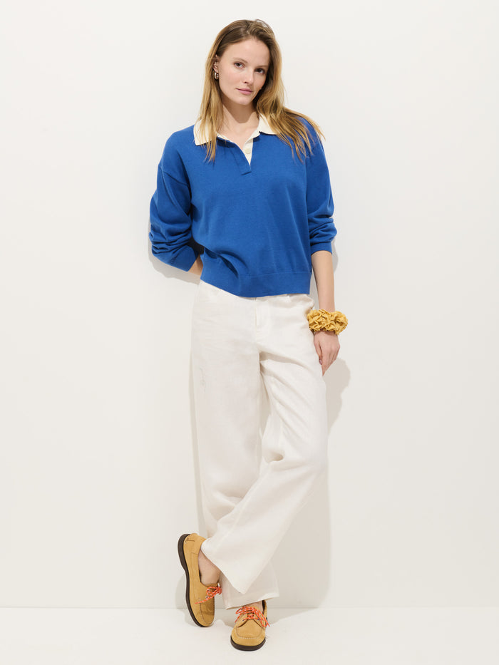 A woman stands against a white background, wearing the Bond St Polo In Cotton Cashmere in rugby blue over a white shirt, paired with white wide-leg pants, tan shoes with orange laces, and a yellow scrunchie on her wrist.