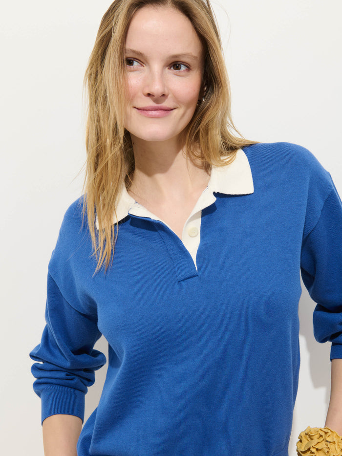 A woman with long blonde hair wears the Bond St Polo In Cotton Cashmere, a relaxed-fit blue sweater with white trim, standing against a plain white background and smiling slightly as she looks to the side.