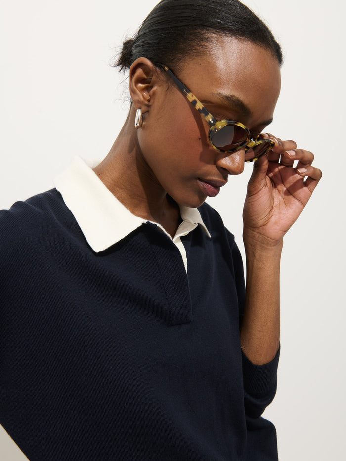 A person in the Bond St Polo In Cotton Cashmere, paired with oval tortoiseshell sunglasses and chunky hoop earrings, looks down while adjusting their sunglasses. Their short hair is pulled back.