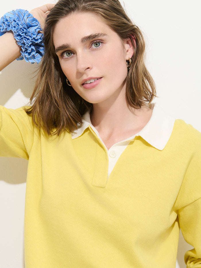 A woman with shoulder-length brown hair wears the Bond St Polo In Cotton Cashmere—an oversized yellow rugby-style shirt with a collared neckline—and a blue scrunchie on her wrist, posing against a white background in bright lighting.