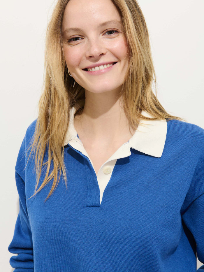 A woman with long blonde hair smiles at the camera, wearing the Bond St Polo in Cotton Cashmere, a relaxed-fit rugby shirt with a white collar and buttons, standing against a plain light background.