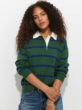 A person with curly hair wears a green and blue Bond St Striped Polo in Cotton Cashmere, styled in a relaxed fit with a white collar and blue jeans, standing against a white background.