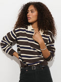 A woman with long curly hair stands against a white background, looking to the side with one hand on her hip. She wears the Bond St. Striped Polo in Cotton Cashmere, featuring relaxed fit brown, cream, and blue stripes, paired with black pants.