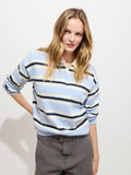 A woman with long blonde hair stands against a plain white background, looking at the camera. She wears gray pants paired with the Bond St. Striped Polo in Cotton Cashmere—a light blue and white striped sweater in an oversized, relaxed fit—adding to her effortlessly stylish appearance.