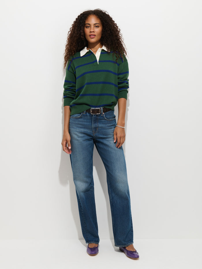 A woman with curly hair wears the Bond St Striped Polo in Cotton Cashmere—featuring green and navy stripes and a relaxed fit—paired with blue jeans, a black belt, and purple flats, standing against a plain white background.