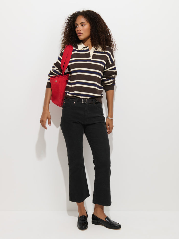 Wearing the Bond St. Striped Polo in Cotton Cashmere, a woman with curly hair stands against a plain white background. She pairs the relaxed-fit long-sleeve top with black pants, black loafers, and carries a red shoulder bag.