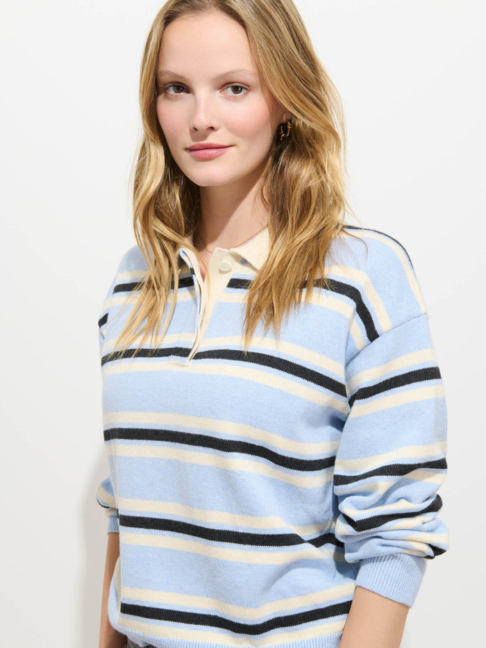 A woman with long blonde hair wears the Bond St. Striped Polo in Cotton Cashmere, featuring light blue, beige, and navy stripes in an oversized fit. She stands against a plain white background, smiling subtly at the camera to showcase the relaxed silhouette and cozy knit of the sweater.