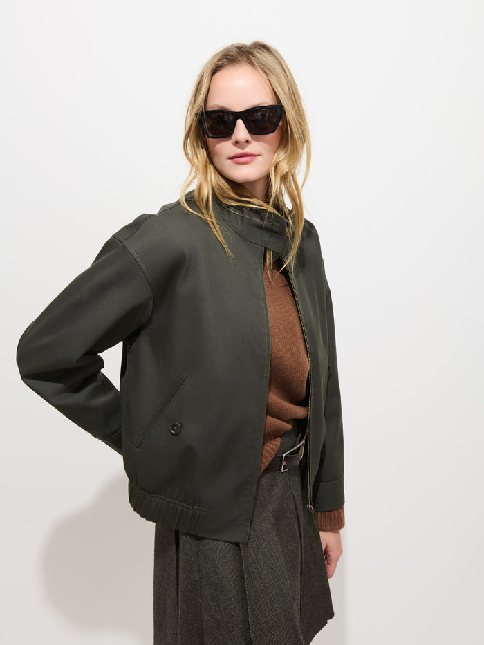 A woman wearing dark sunglasses, the Brighton Zip Jacket in Twill Gabardine—a dark green jacket crafted from luxurious Olmetex Italian Gabardine—a brown sweater, and a grey skirt poses against a plain white background with one hand on her hip.