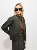 A woman wearing dark sunglasses, the Brighton Zip Jacket in Twill Gabardine—a dark green jacket crafted from luxurious Olmetex Italian Gabardine—a brown sweater, and a grey skirt poses against a plain white background with one hand on her hip.