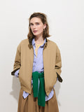 A woman stands against a white background wearing the Brighton Zip Jacket in Twill Gabardine, styled with a blue striped shirt, tan skirt, and a green sweater tied around her waist. She has light brown hair and a relaxed expression, with her hands in her jacket pockets.