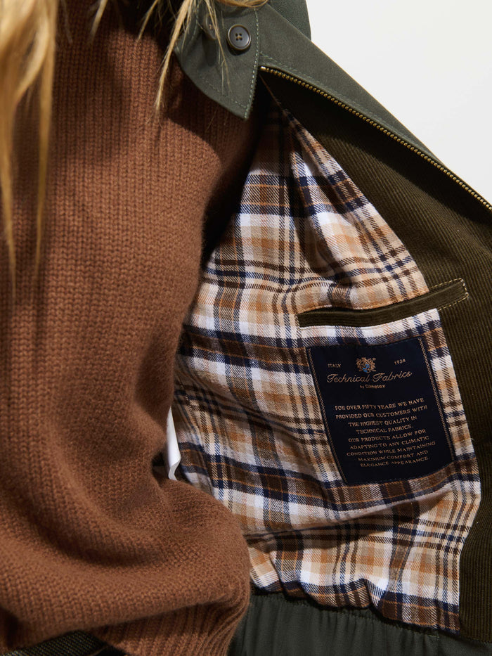 A person wearing a brown sweater holds open the Brighton Zip Jacket in Twill Gabardine in olive green, revealing its brown plaid lining and an interior label that reads Technical Fabric. Crafted from Olmetex Italian gabardine, the jacket features water-repellent fabric for added protection and style.
