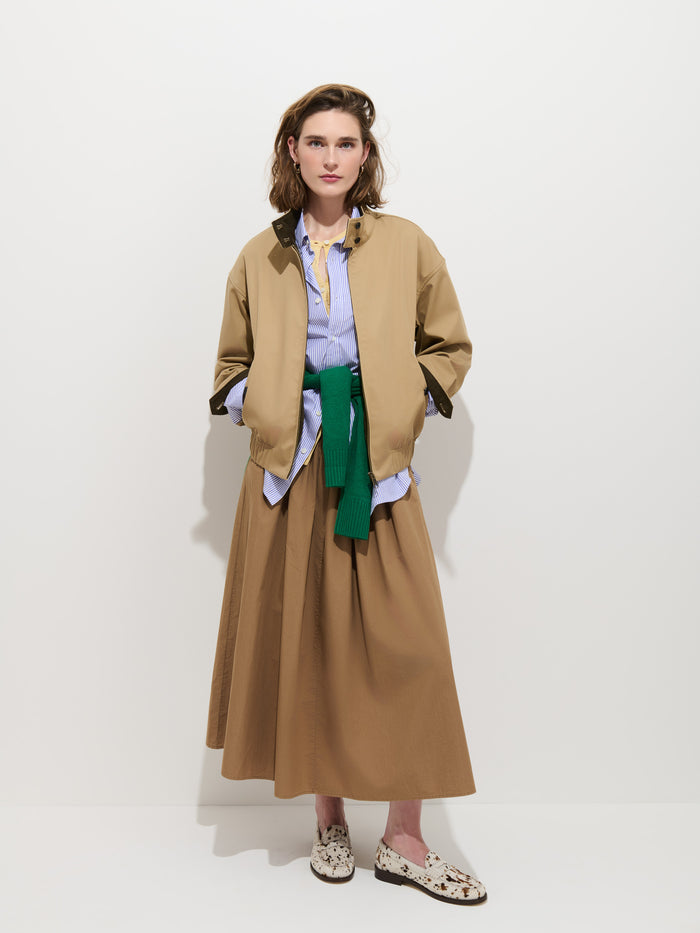 A woman stands against a plain white background wearing the Brighton Zip Jacket in Twill Gabardine, paired with a blue shirt, tan skirt, and loafers. A green sweater is tied around her waist as she looks at the camera with a neutral expression.