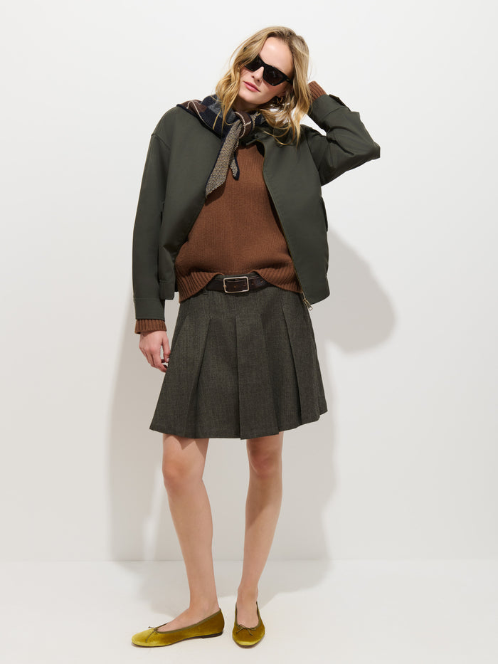 A woman stands against a white wall, wearing sunglasses and the Brighton Zip Jacket in Twill Gabardine—crafted from olive water-repellent fabric—paired with a brown sweater, pleated skirt, yellow flats, and a scarf around her neck; she raises one arm to touch her head.