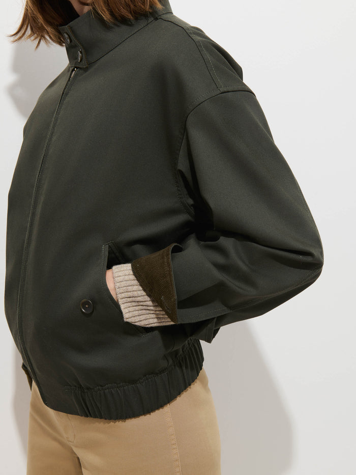 A person wears the Brighton Zip Jacket in Twill Gabardine, a dark green, water-repellent jacket crafted from Italian Gabardine, paired with beige pants. They stand against a plain white background with one hand in the jacket pocket and a striped sleeve partially visible.