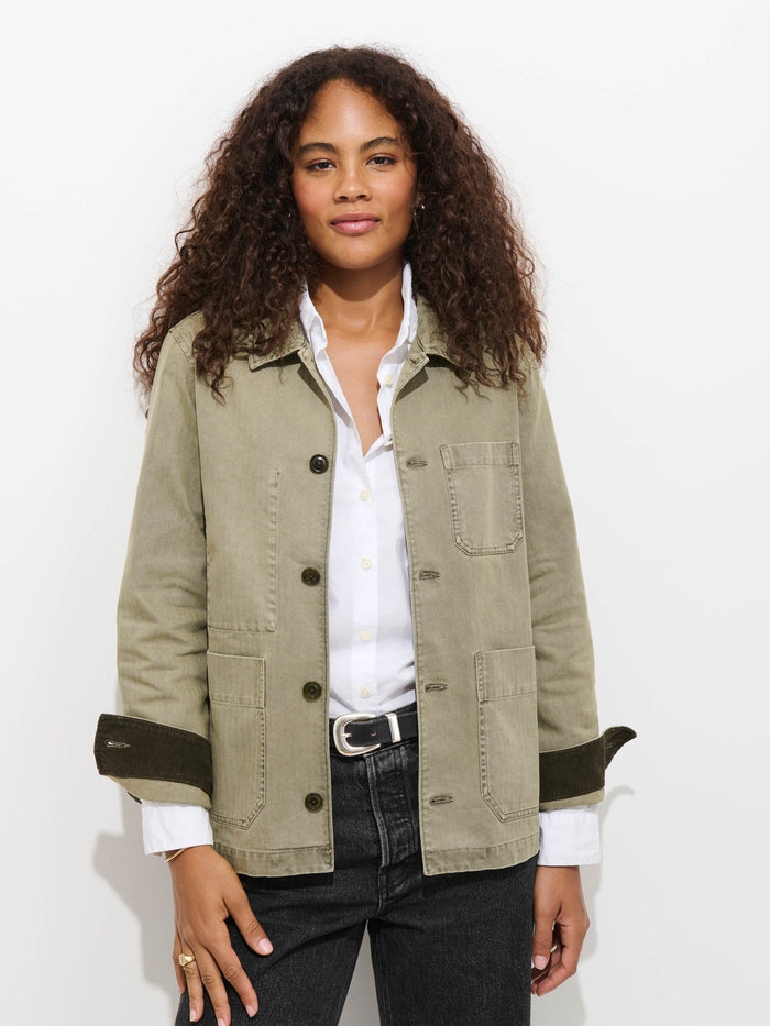 A person with curly hair wears the Britt Work Jacket In Herringbone, which features a corduroy collar, over a white button-up shirt and dark jeans, standing against a white background with a relaxed expression.