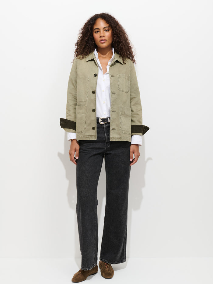 A woman with curly hair stands against a white background wearing the Britt Work Jacket in Herringbone over a white button-down shirt, paired with dark wide-leg jeans, a black belt, and brown loafers.